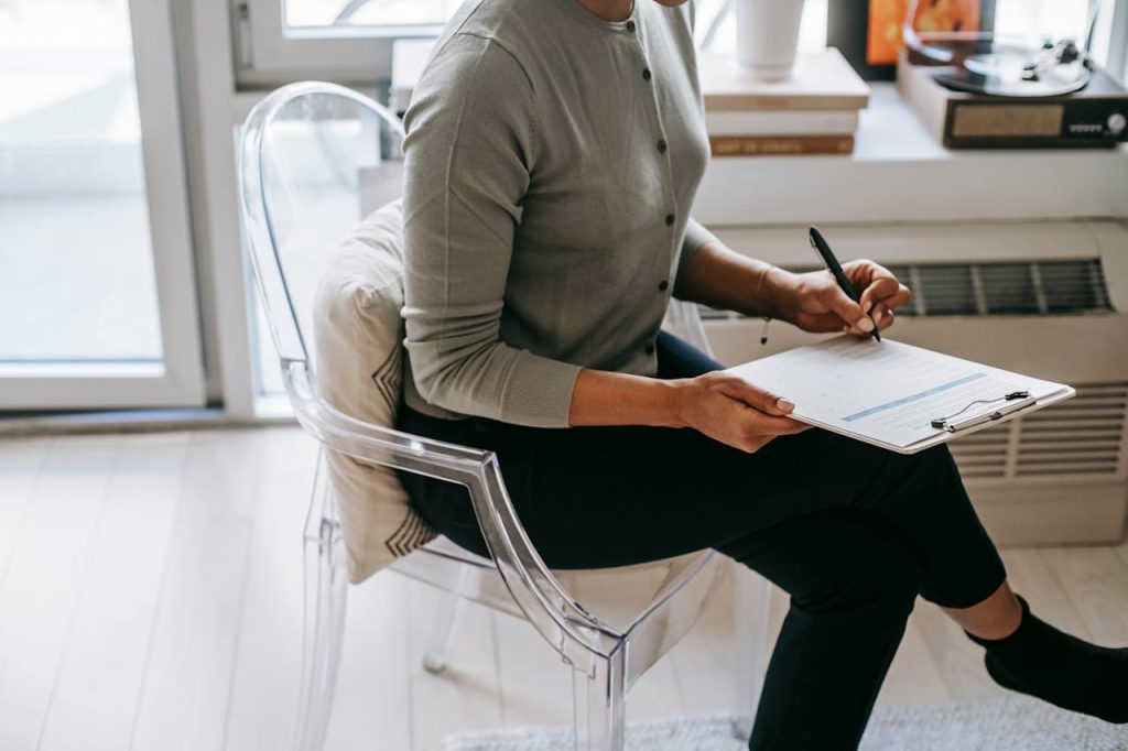 From above of unrecognizable female psychologist with crossed legs taking notes on clipboard while filling out questionnaire during psychotherapy appointment in cozy office